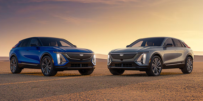 A blue and a silver Cadillac Lyriq electric SUV parked side by side in a desert landscape during sunset.