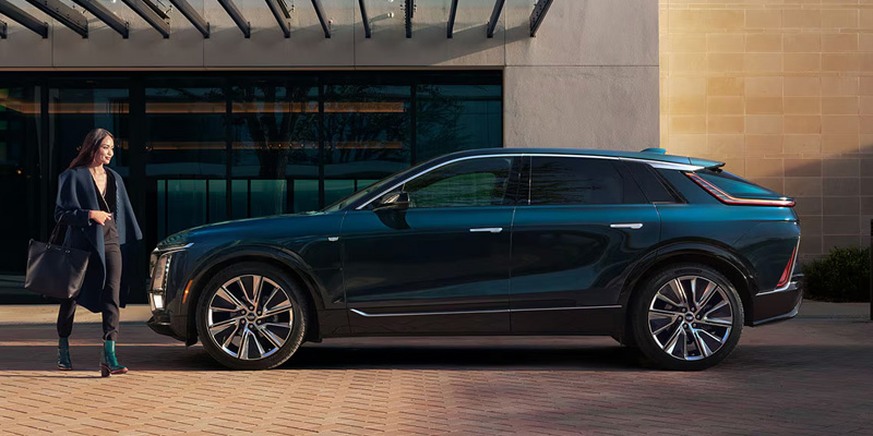 A deep teal Cadillac LYRIQ parked in front of a modern building as a stylish woman walks toward the vehicle.