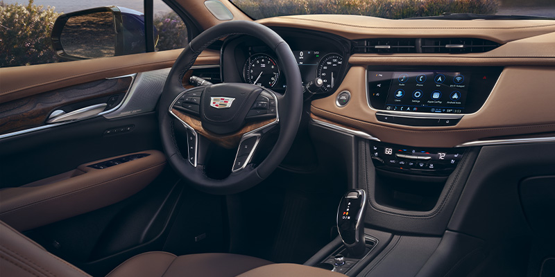 he luxurious interior of a Cadillac featuring a tan and black leather dashboard, wood grain accents on the steering wheel and door panels, and a large central infotainment touchscreen displaying various app icons.