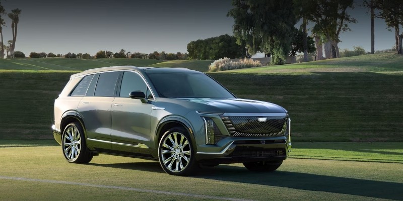 Front-three-quarter view of a metallic gray Cadillac VISTIQ SUV parked on a manicured green golf course fairway with palm trees and rolling hills in the background.