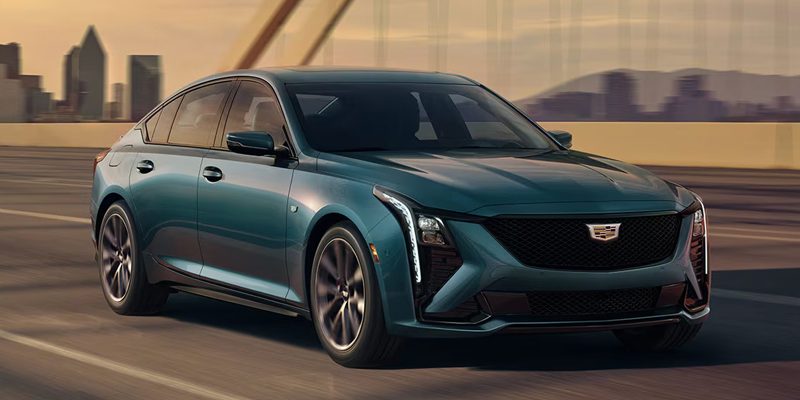 A teal blue Cadillac CT5 sedan driving along a bridge with a city skyline at sunset.