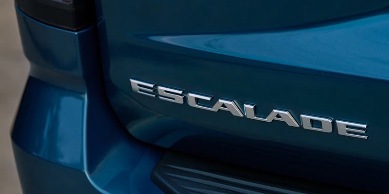 A close-up shot of the silver chrome "ESCALADE" emblem mounted on the teal blue body of a Cadillac SUV.