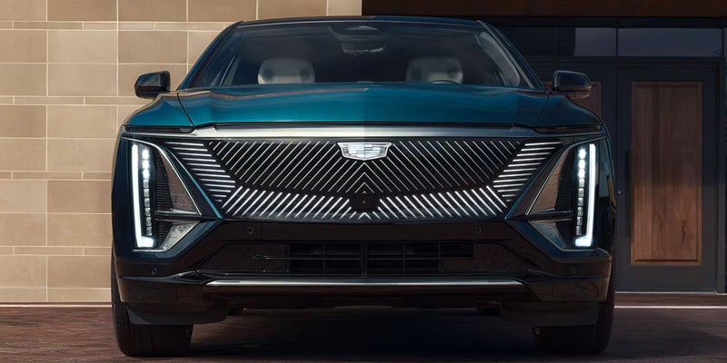 Front head-on view of a dark teal Cadillac Lyriq EV showing its illuminated black crystal shield grille and vertical LED light blades.