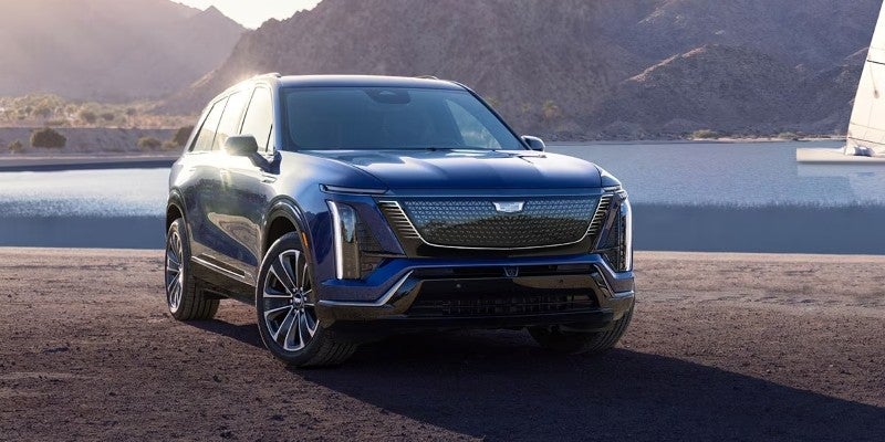 A dark blue Cadillac Escalade IQ electric SUV parked near a body of water with mountains in the background.